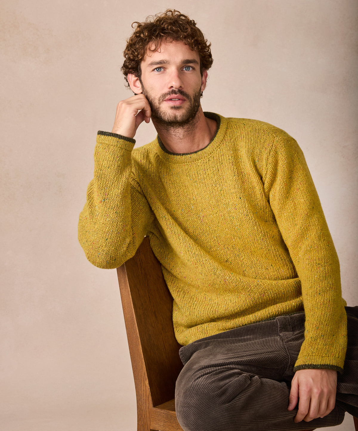 Man wearing a mustard yellow sweater sitting on a wooden chair against a beige background