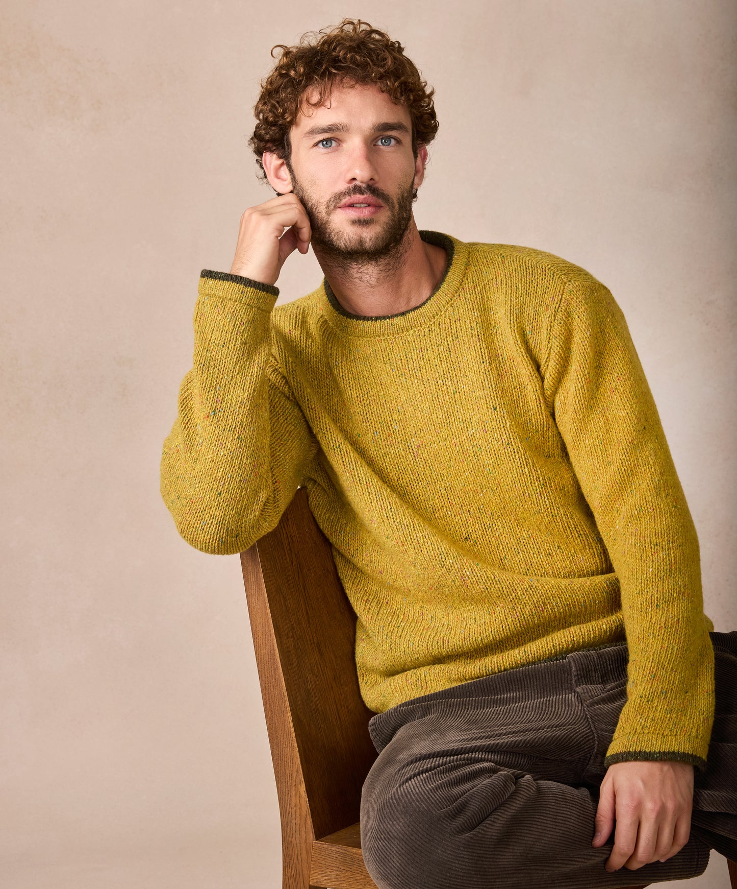 Man wearing a mustard yellow sweater sitting on a wooden chair against a beige background