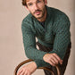 Man wearing a green knitted sweater sitting on a wooden chair against a beige background