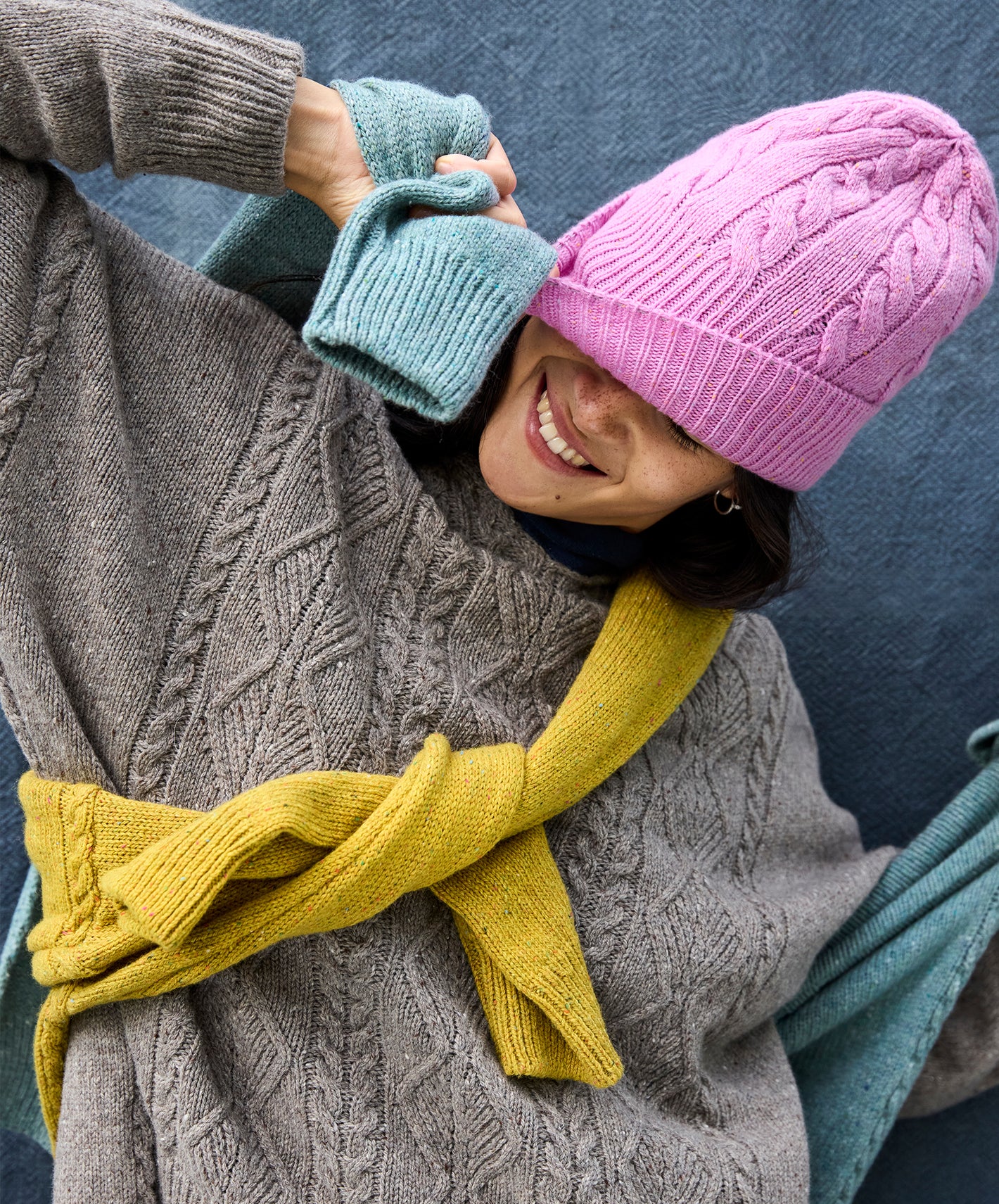 Person wearing an IrelandsEye pink knit elio hat, grey ada hoodie, yellow isla sweater, and blue isla sweater against a dark background.